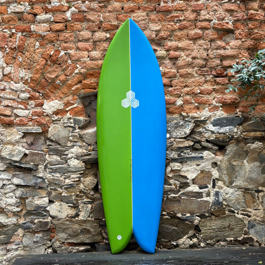 Green and blue CHANNEL ISLANDS   FEB'S FISH      5’7  	20 1/2	  2 7/16	  33.8L surfboard leaning against a stone wall.