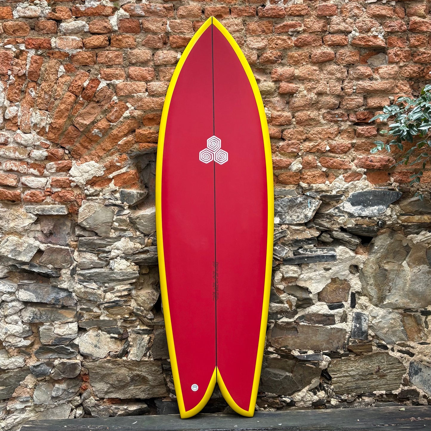 Red CHANNEL ISLANDS FEB'S FISH 5’9 20 7/8 2 1/2 36.3L surfboard with yellow edges against a stone wall
