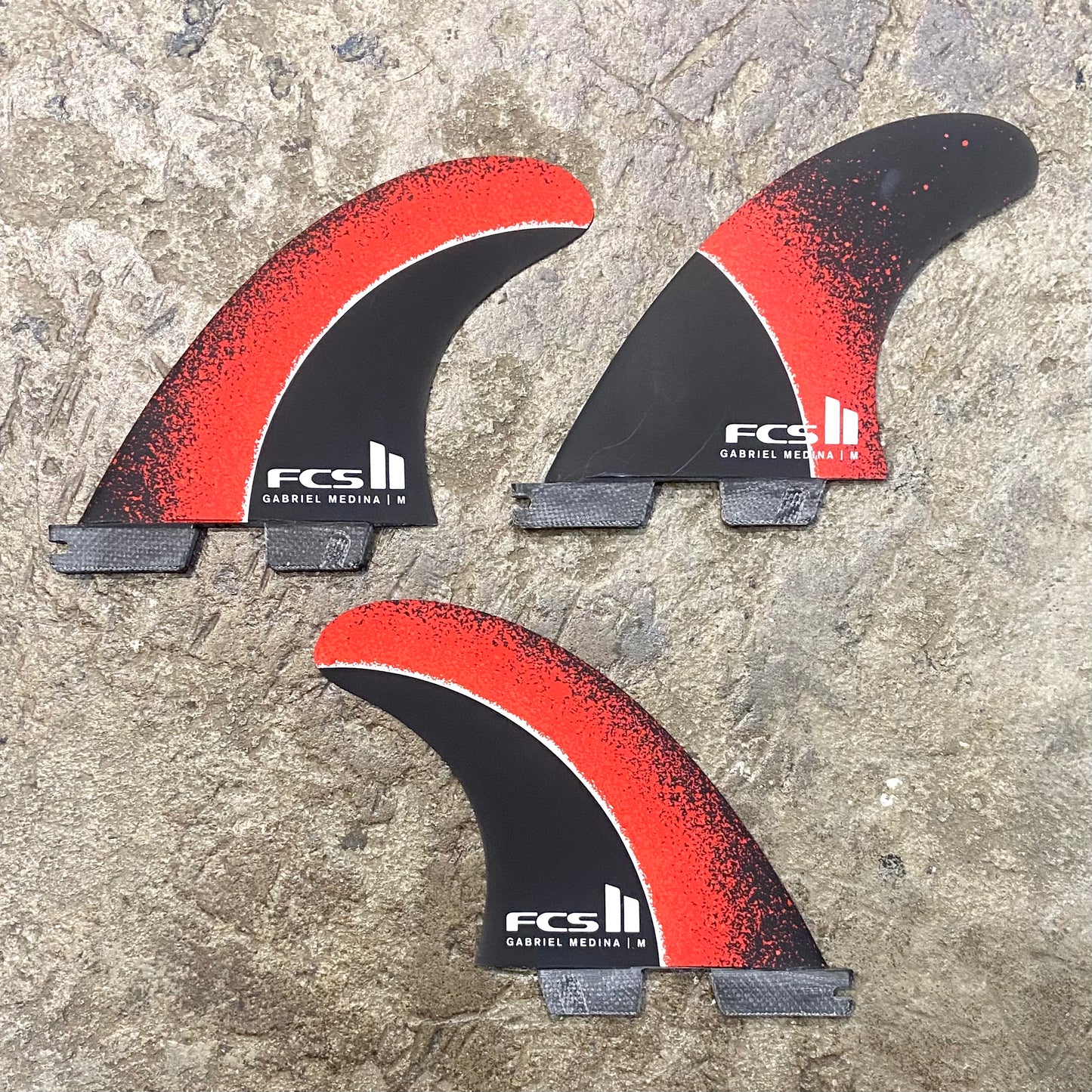 A set of FCS II Gabriel Medina Signature Tri Fins with red, black, and grey color scheme. Laid on concrete floor