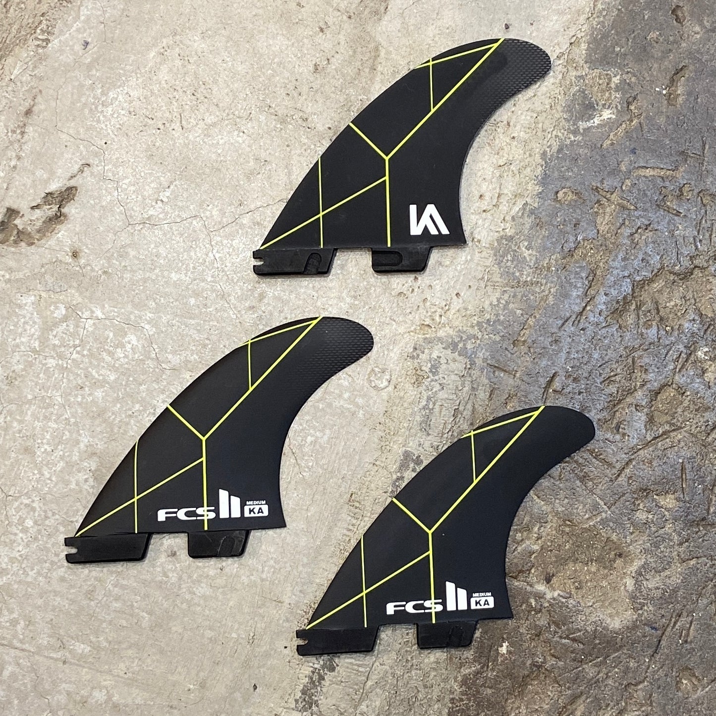 A set of medium FCS II Kolohe Andino PC Tri fin in a black colour way with green details on a concrete floor
