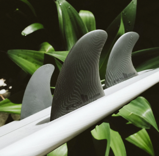 View of the FCS II Reactor Neo Glass Eco Tri Fins, in an all grey colour way in a board with a plant as the background