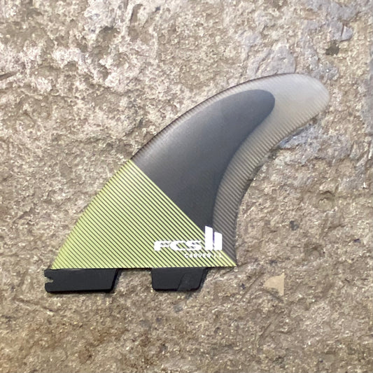A single large fin from a set of FCS ll Essential Series Carver PC Tri Fins in a black and green colour way laid on a concrete surface