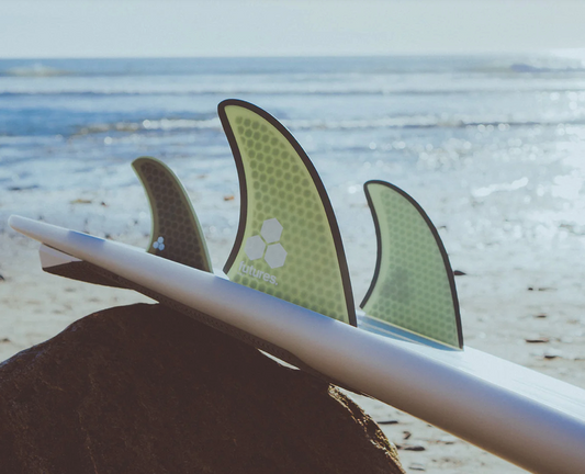 A set of green Futures Al Merrick Twin+1 surfboard fins displayed in a surfboard leaning on a rock at the beach