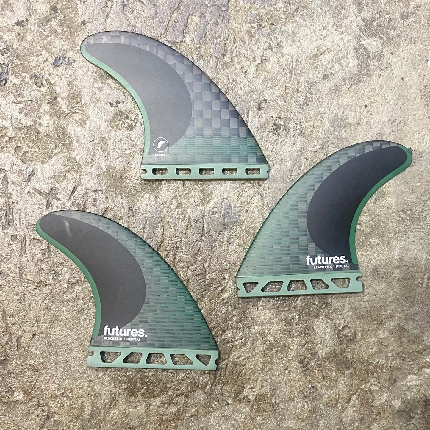 A set of Futures F8 Blackstix black and green surfboard fins with the futures brand logo on them, displayed on a textured grey surface.
