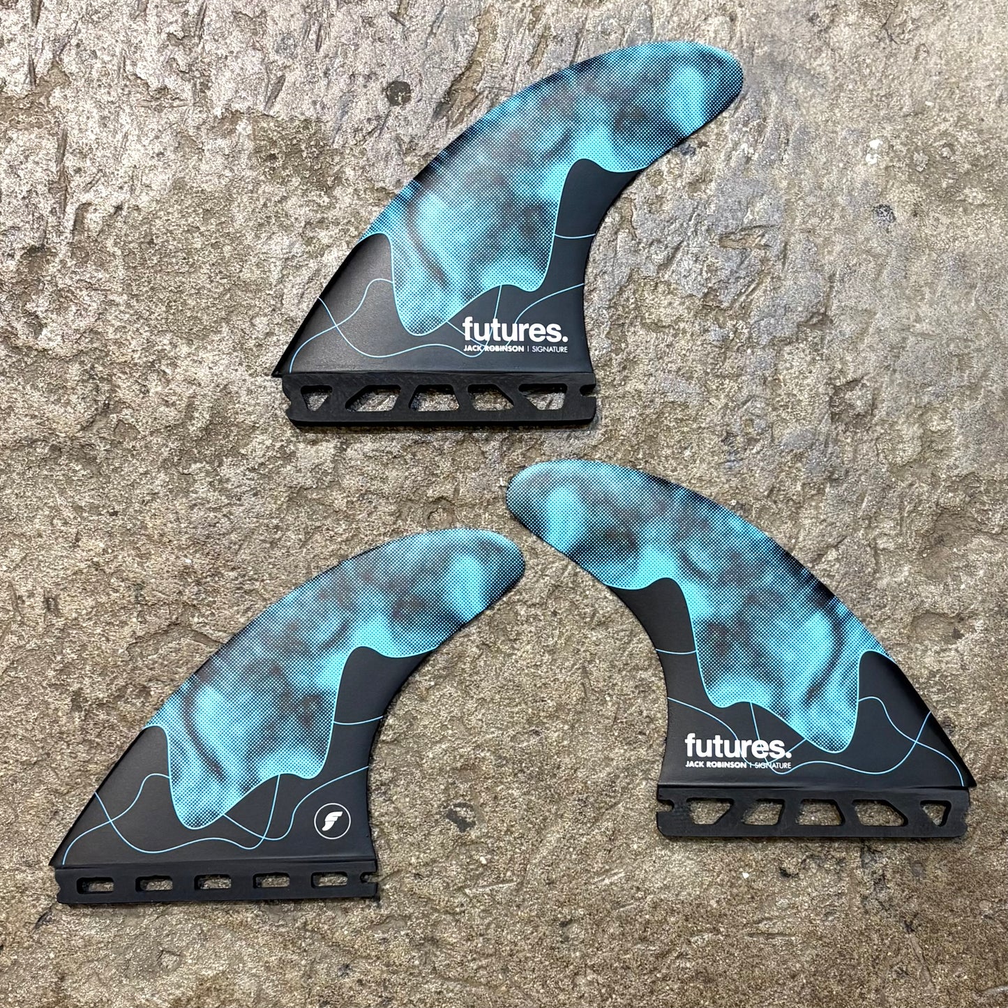 A set of Jack Robinson signature Futures surfboard fins with a honeycomb design, predominantly black with blue accents, laid on a concrete surface.
