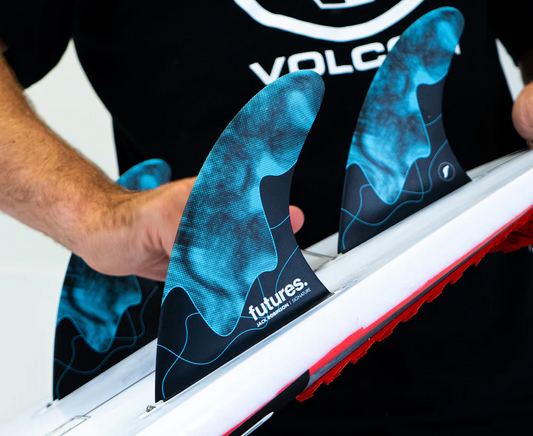 A man screwing a set of Jack Robinson signature Futures surfboard fins into a board. predominantly black colour way with blue accents