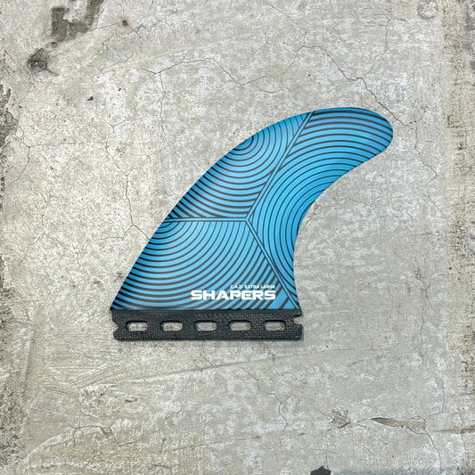 A blue and black surfboard fin with a geometric wave pattern and the brand name 'SHAPERS' printed on it, placed on a textured concrete surface.