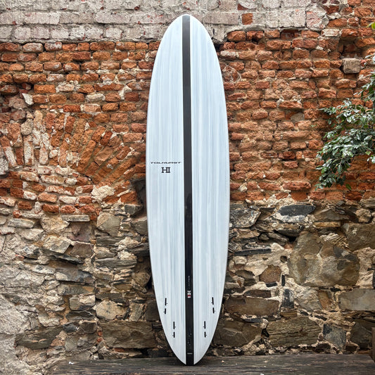 A long surfboard with a white and light gray woodgrain pattern and a black center stripe, leaning against a rustic brick and stone wall.