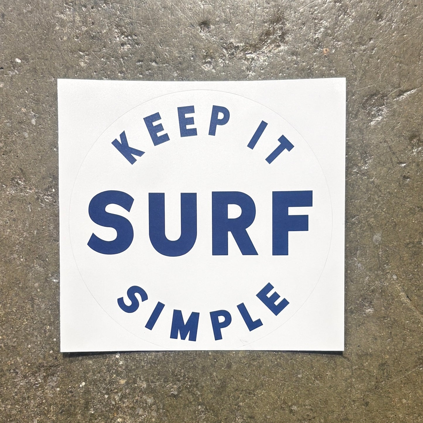 A square white sticker with the words 'KEEP IT SIMPLE SURF' in blue lettering, placed on a textured gray concrete surface.