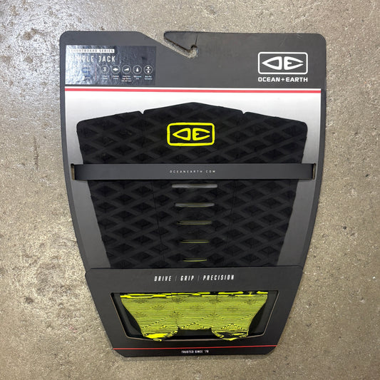 Ocean Earth Simple Jack Traction Deck in its original retail packaging, featuring a black textured grip pad with a yellow logo and a clear window showing the yellow and black grip tape underneath, set against a concrete floor background.