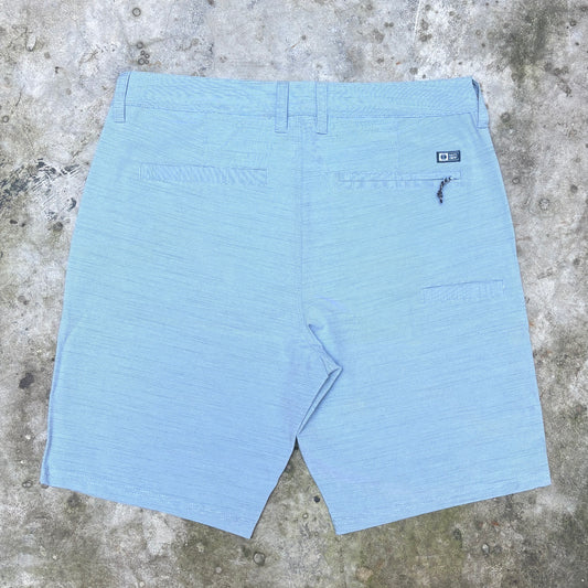 Light blue textured shorts shown from the back, laid flat on a concrete surface. The shorts feature belt loops, a button closure, two back welt pockets, and a small black brand patch on the upper right side.