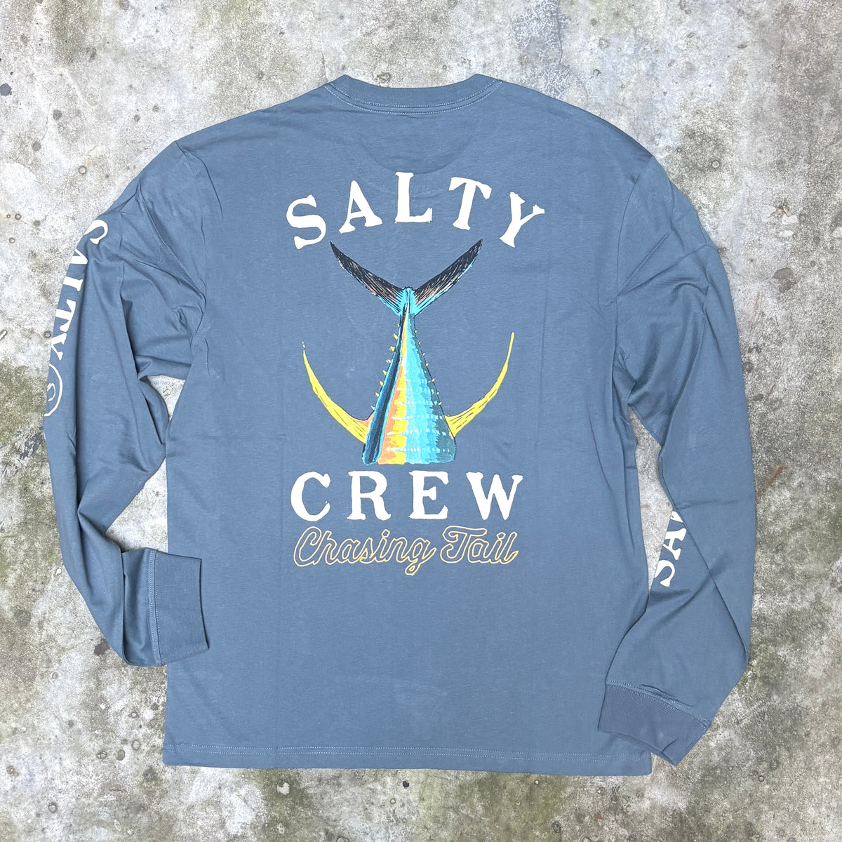 A long-sleeve t-shirt laid flat on a textured concrete background, showing the back graphic. The shirt is slate blue with a large graphic of a blue and yellow marlin tail and the text 'SALTY CREW Chasing Tail'.