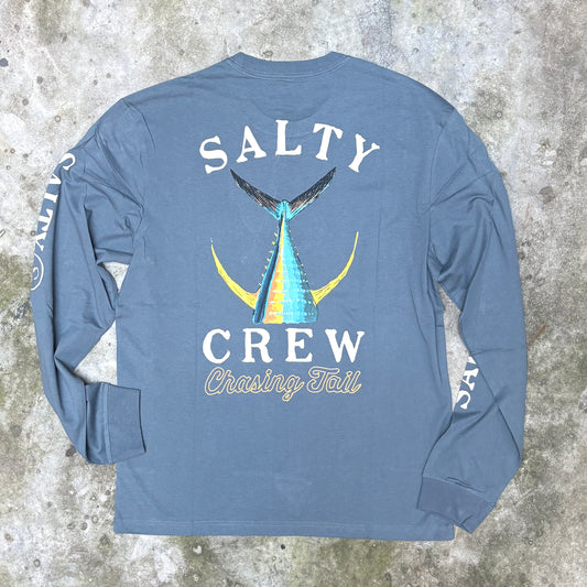 A long-sleeve t-shirt laid flat on a textured concrete background, showing the back graphic. The shirt is slate blue with a large graphic of a blue and yellow marlin tail and the text 'SALTY CREW Chasing Tail'.