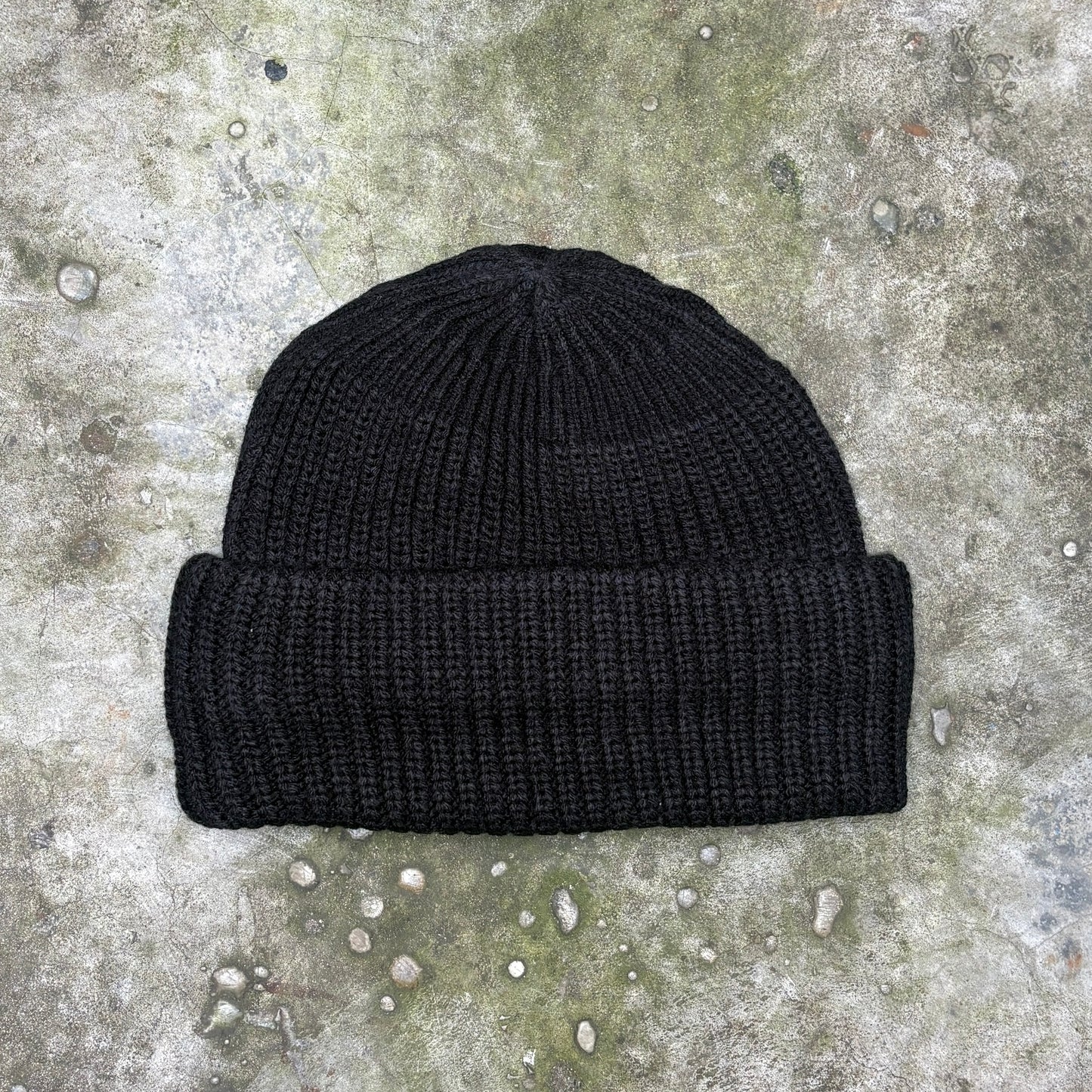 A black ribbed knit beanie hat laid flat on a textured concrete surface.