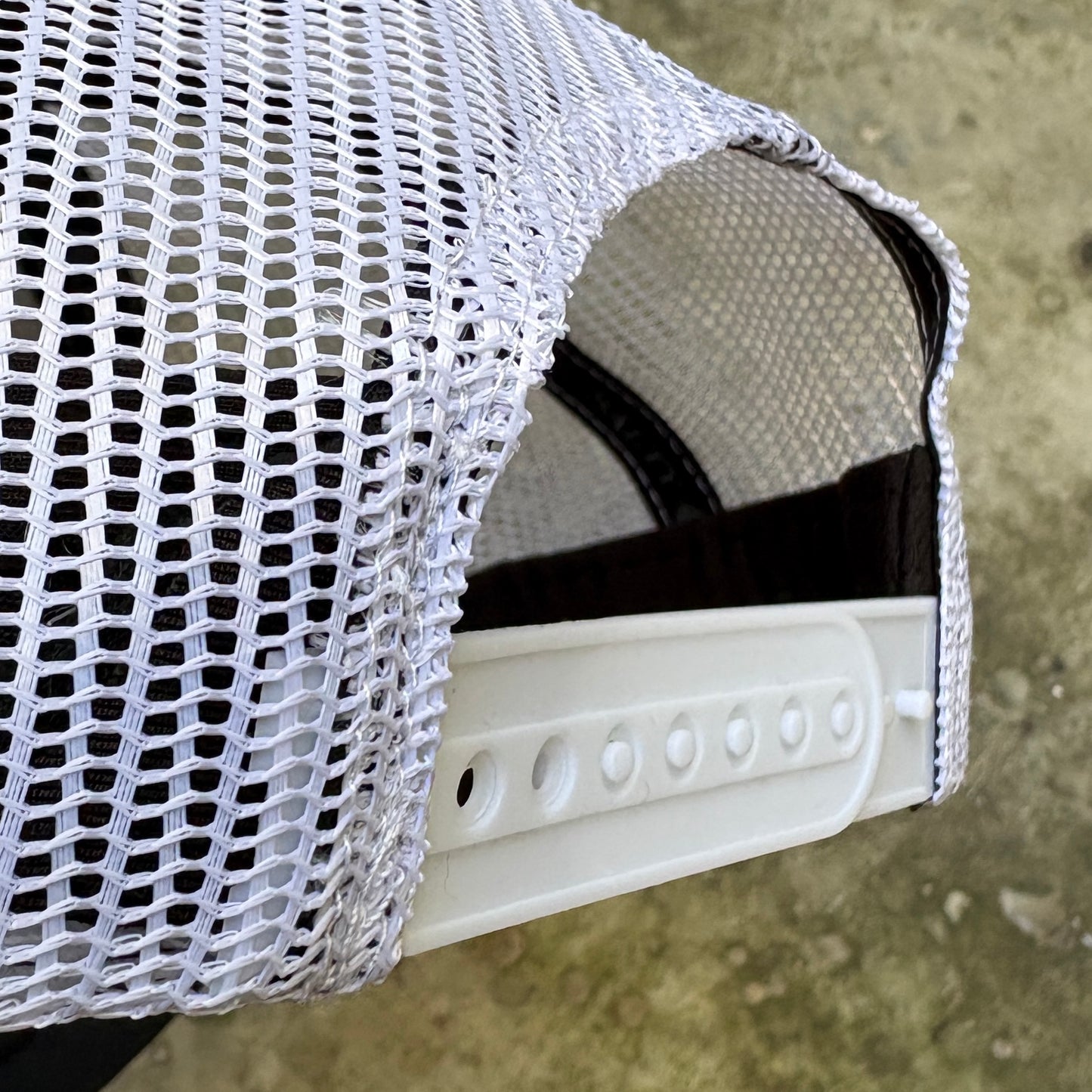 Close-up of a white mesh trucker hat showing the side and back panel with adjustable ventilation holes and a white plastic visor.