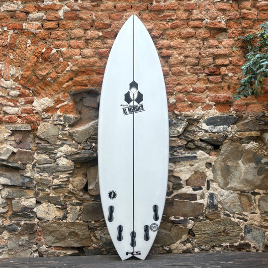 A white Channel Islands surfboard with black graphics and fin hardware, standing upright against a rustic brick and stone wall.