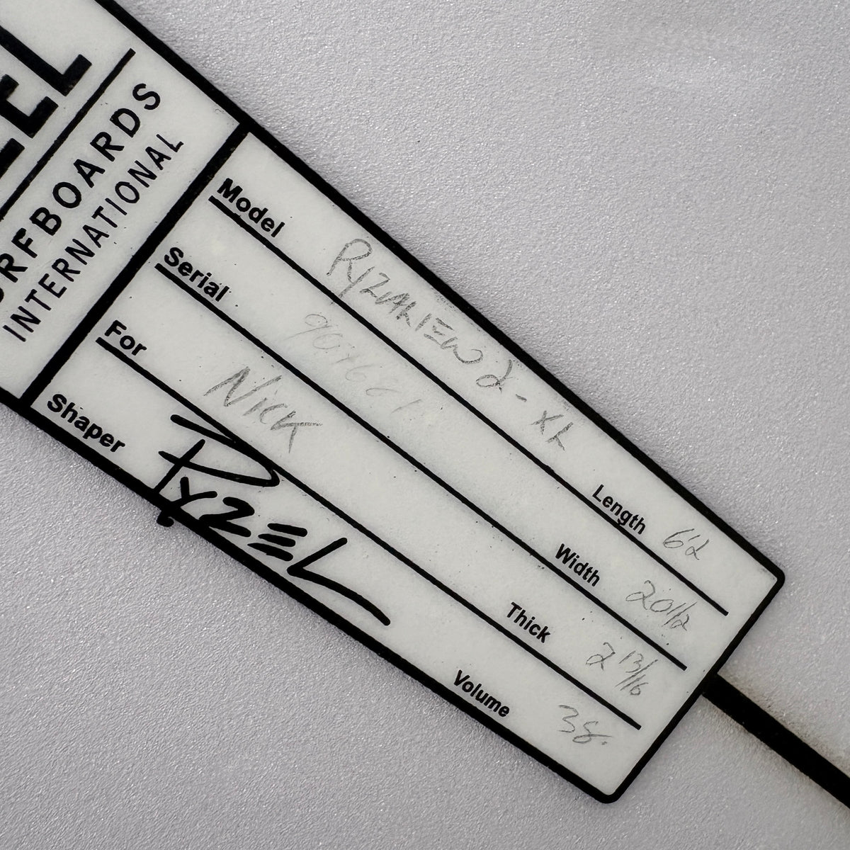 A close-up of a white and black custom surfboard maker's production card with handwritten details, placed on a textured white background.