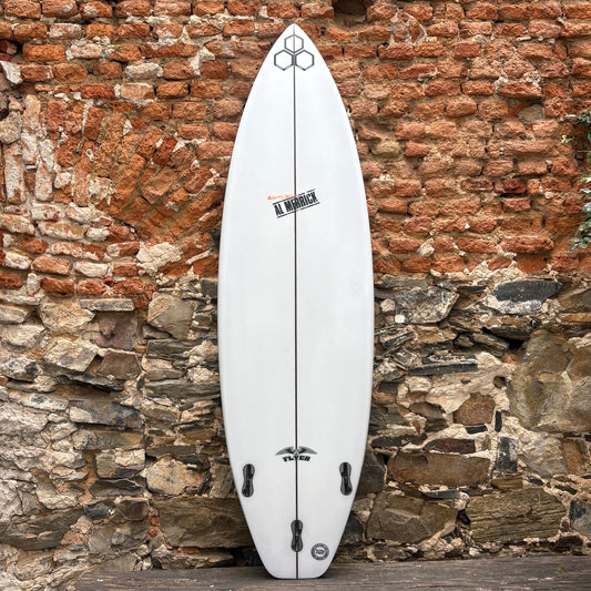 A white surfboard with black graphics and hardware, standing upright against a rustic brick and stone wall.