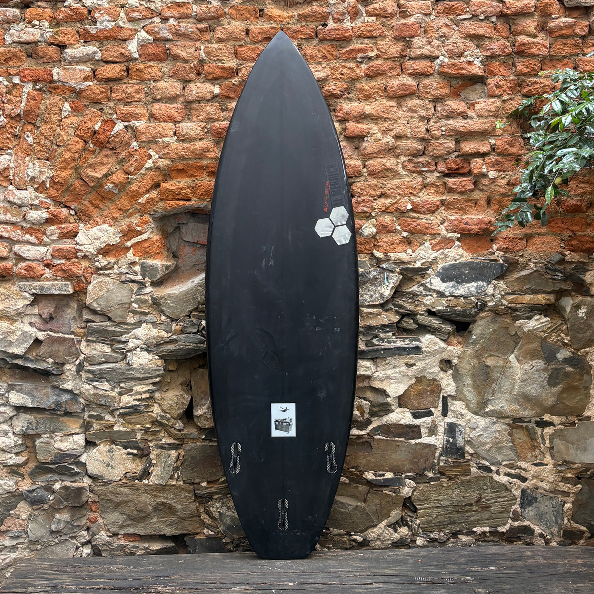 A black surfboard standing upright against a rustic stone and brick wall.