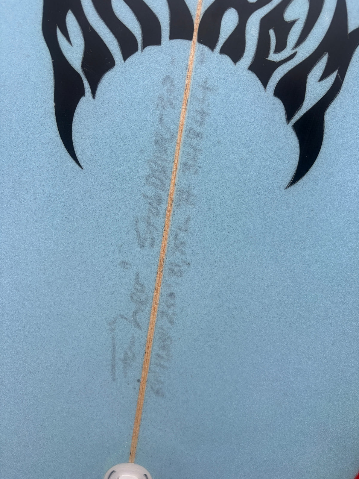Close-up of a light blue surfboard with a black flame graphic at the top and a visible center split line, photographed without a person or real environment.