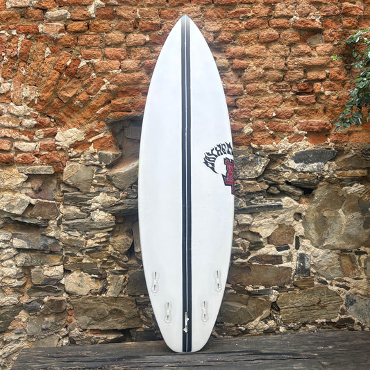 A white surfboard with a black center stripe and a black-and-red 'Milk Cow' logo near the tail, leaning against a rustic brick and stone wall.