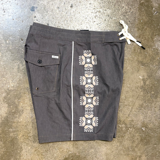 Dark gray board shorts with a vertical beige and white geometric pattern down the center, laid flat on a concrete floor.