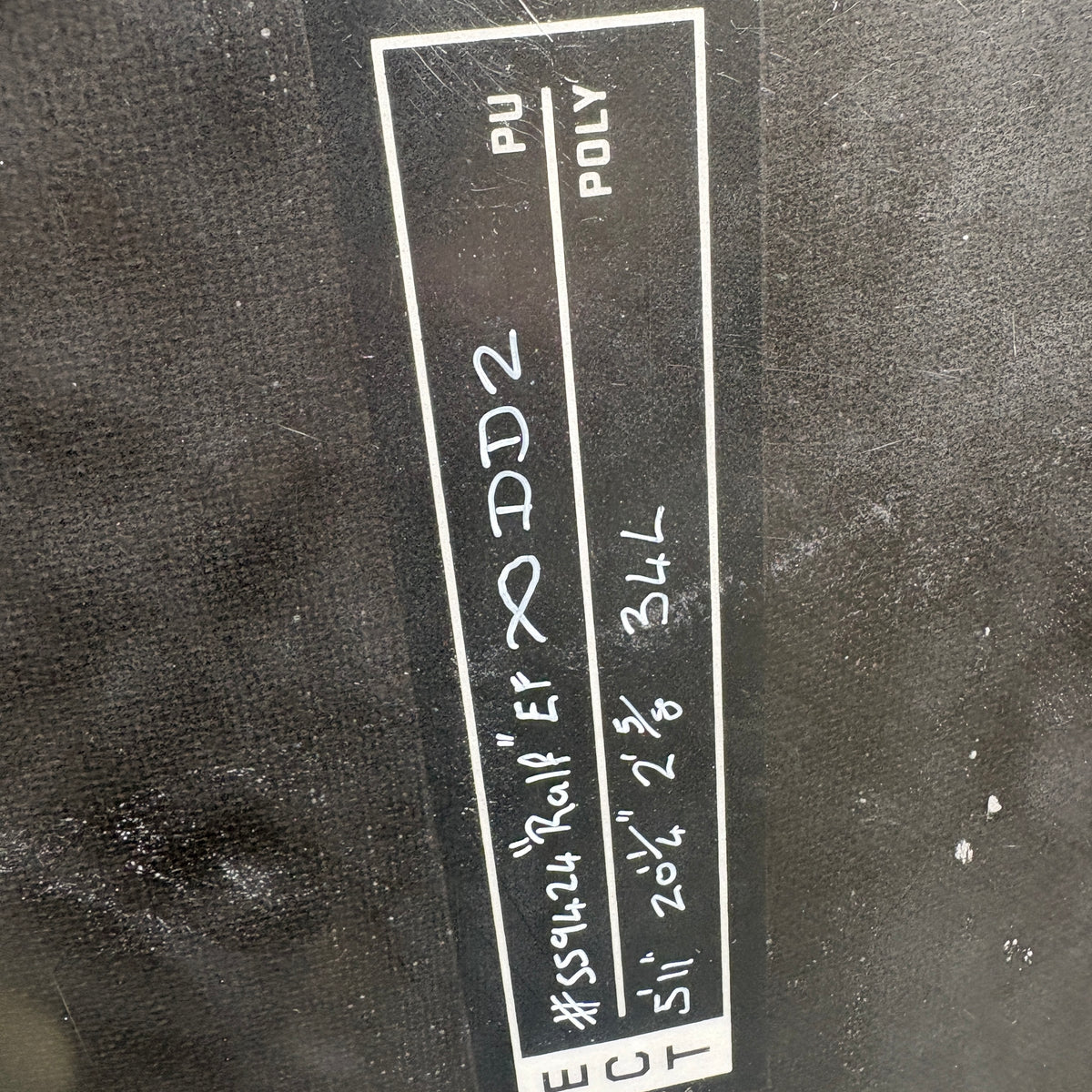 A close-up of a black fabric label with white text and handwritten markings, attached to a dark textured garment or material.