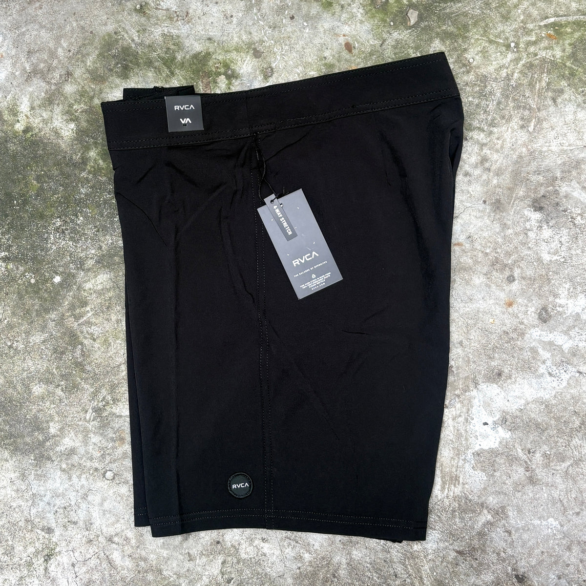 Black RVCA athletic shorts laid flat on a textured concrete surface, with a size tag, product tag, and small circular logo patch visible.