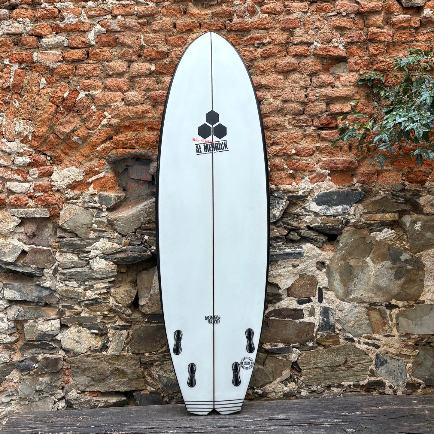 White Bobby Quad surfboard with black logo against a rustic stone wall