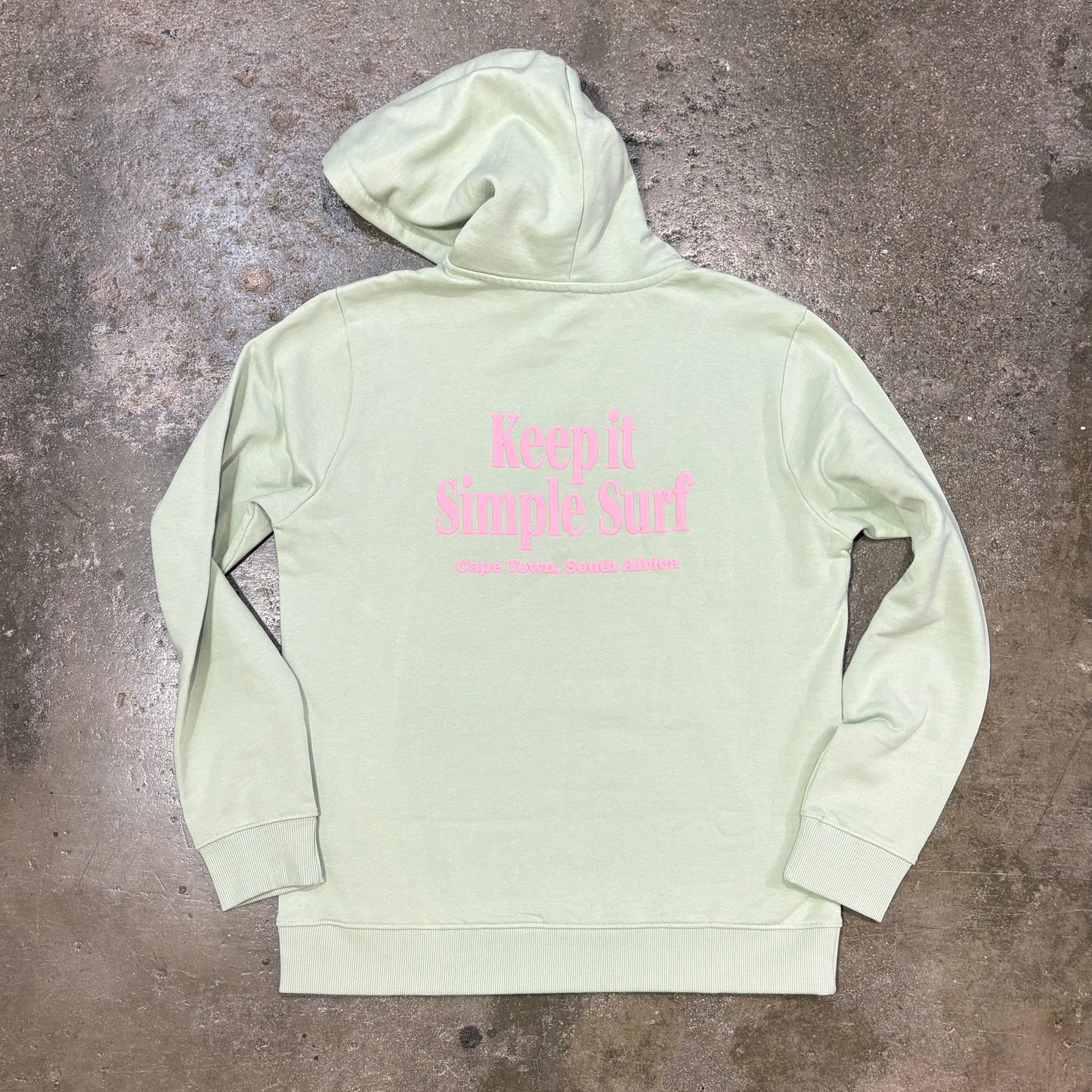 A light sage green hoodie with pink text reading 'Keep It Simple Surf' on the back, laid flat on a textured concrete floor.