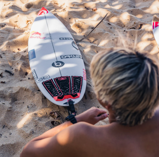 Person sitting on a sandy beach with a surfboard featuring Oakley sticker and Ocean&Earth Surf Traction