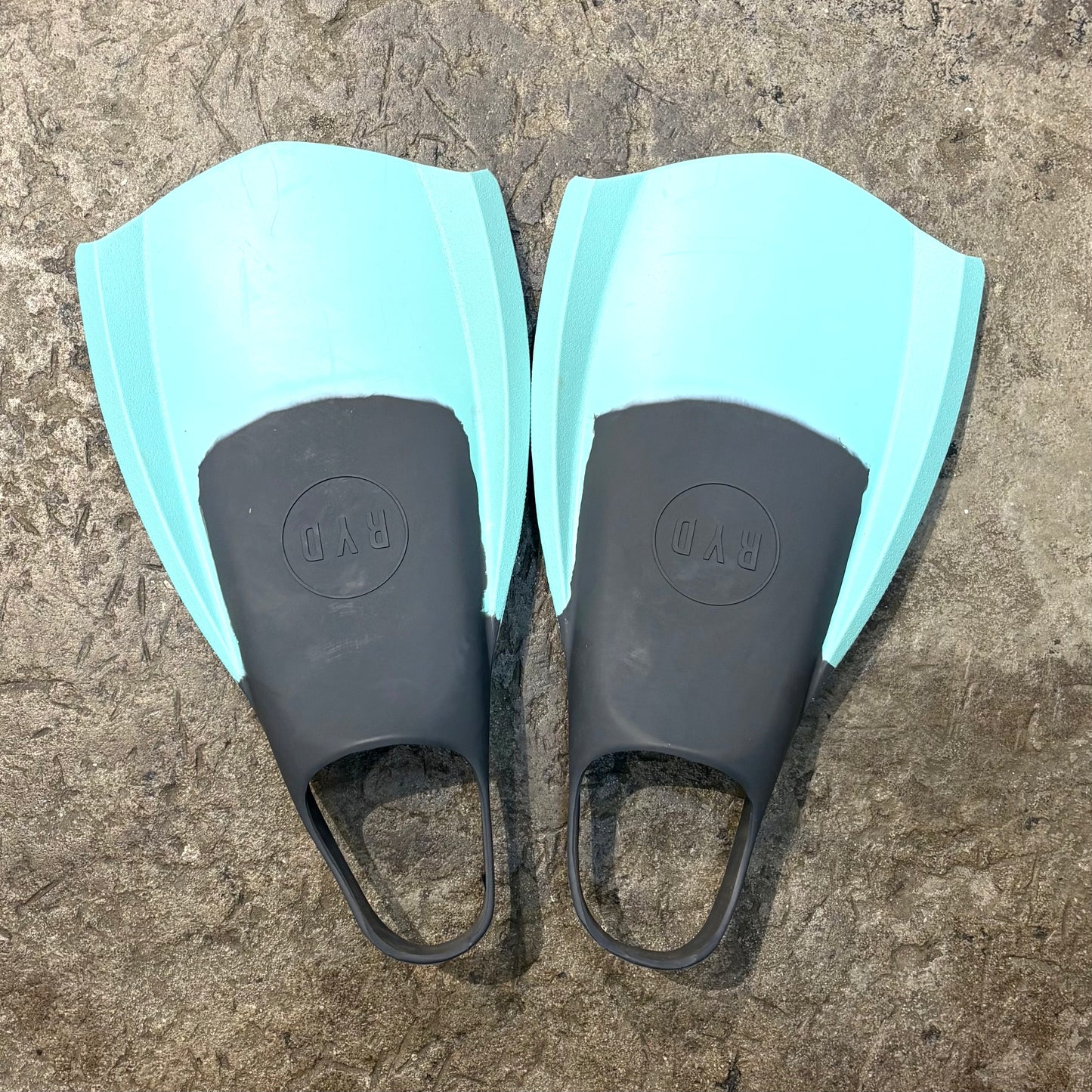 Pair of swim fins with black blades and light blue straps on a textured surface