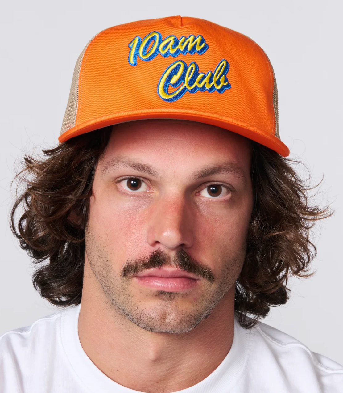 A man with wavy brown hair and a mustache wearing an orange trucker hat with '10am Club' embroidered on the front, photographed against a light gray background.