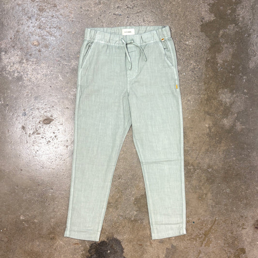 A pair of light sage green drawstring linen trousers laid flat on a textured concrete floor.