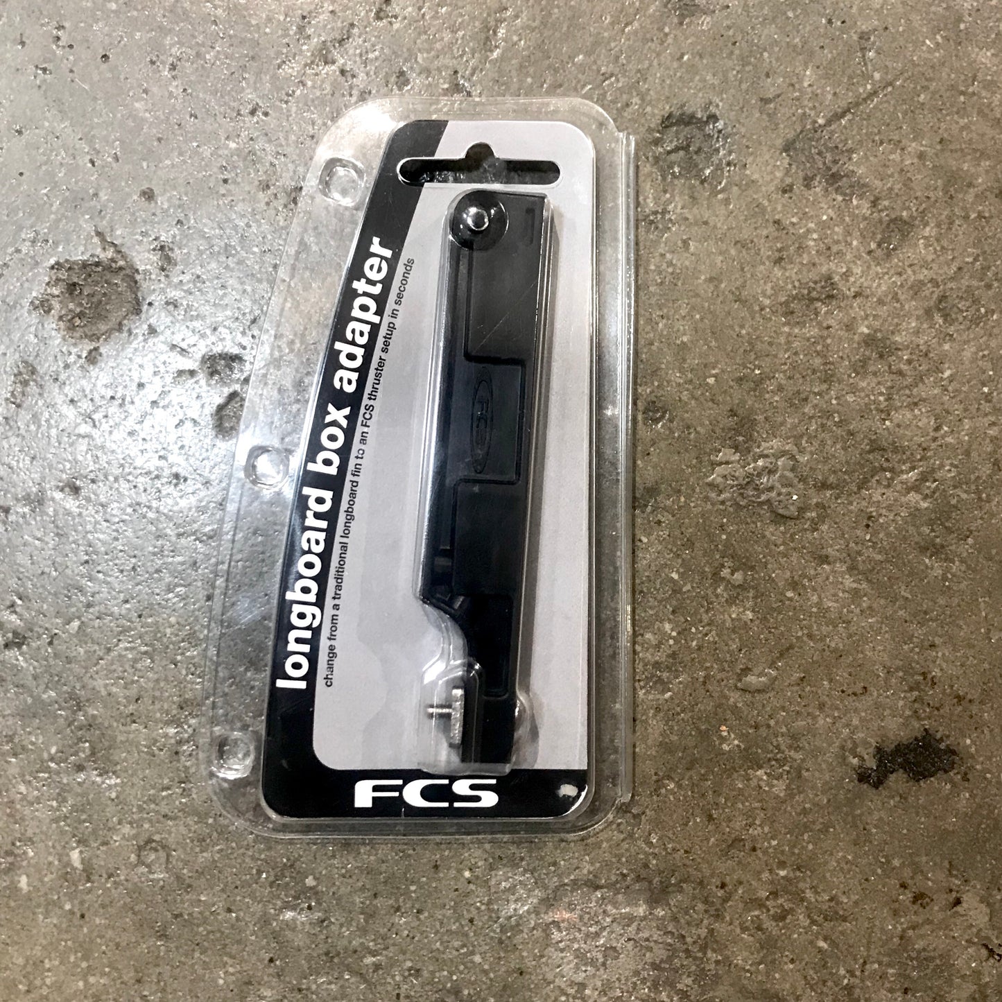 A FCS Longboard Box Adapter in it's packaging laid out on a concrete surface