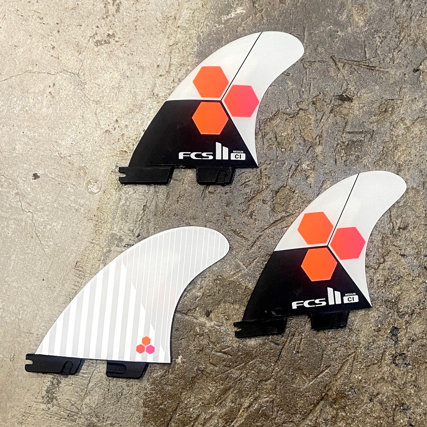 View of a set of FCS ll CI Upright Tri Fins in a white, black and orange colourway laid out on a concrete floor.
