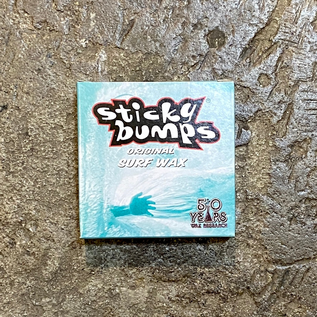 A small rectangular surfboard wax package with 'Sticky Bumps Original Surf Wax' text and a blue and white design, placed on a textured surface.
