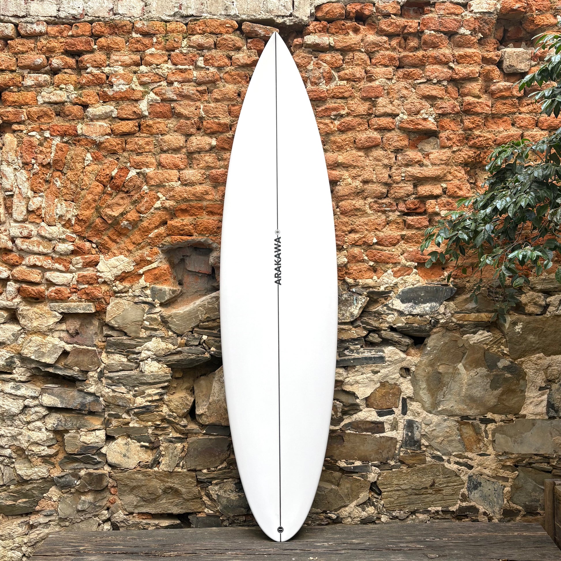 White surfboard with 'Arakawa' branding against a rustic stone wall.