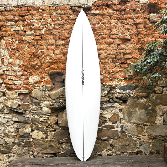 White surfboard with 'Arakawa' branding against a rustic stone wall.
