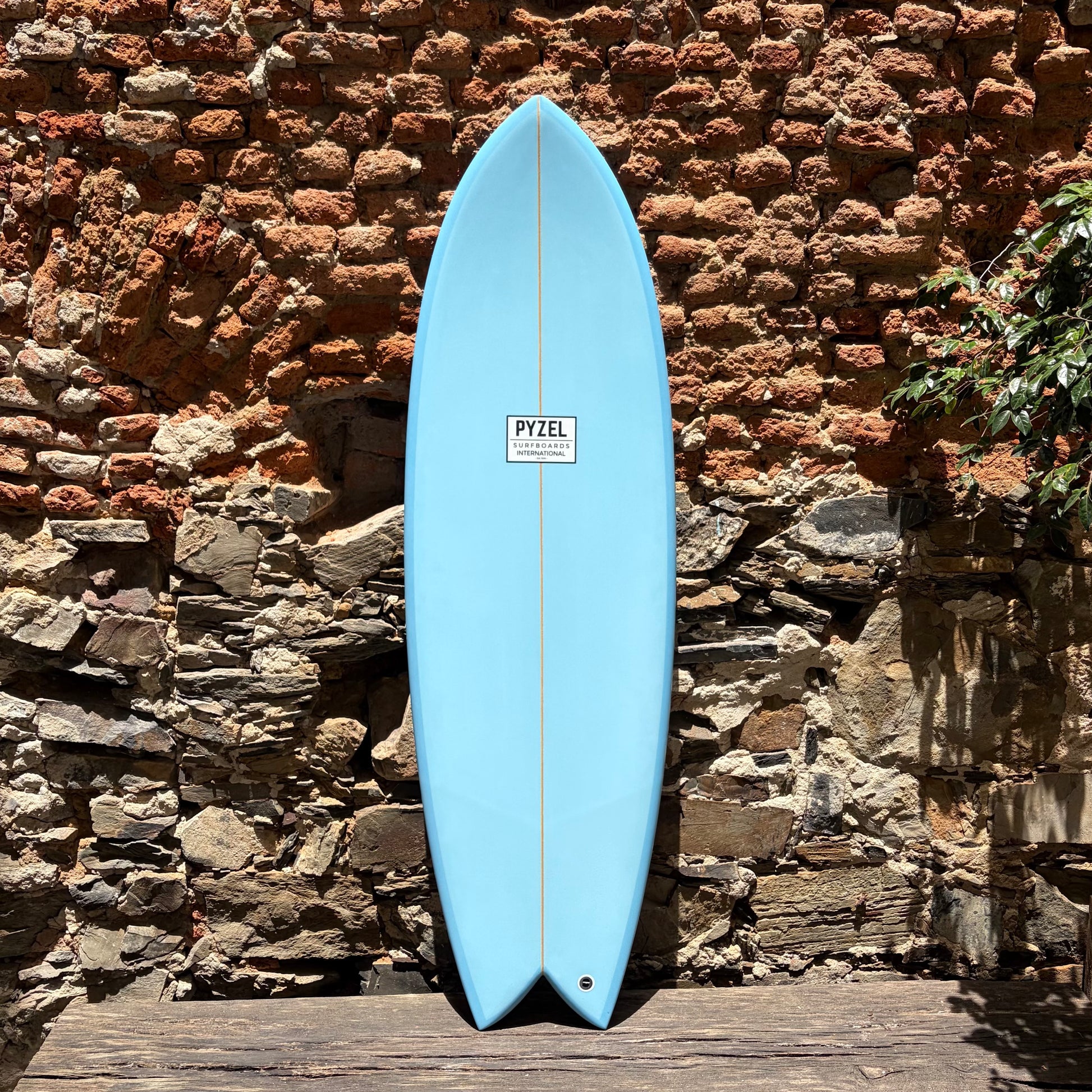 Blue Pyzel surfboard with a brown stripe against a rustic brick wall.