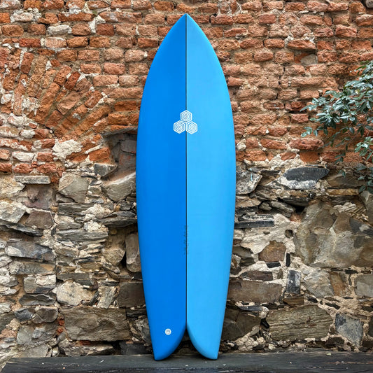 Blue CHANNEL ISLANDS   FEB'S FISH      5'11   21 1/8	  2 5/8	  39.7L surfboard with a logo against a stone wall