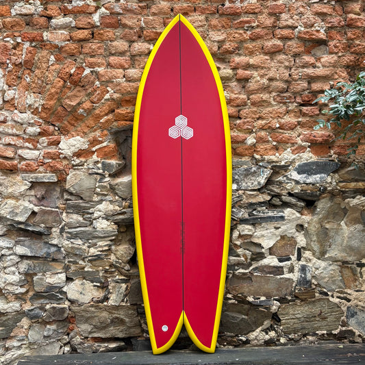 Red CHANNEL ISLANDS   FEB'S FISH      5’9    20 7/8	  2 1/2	  36.3L surfboard with yellow edges against a stone wall