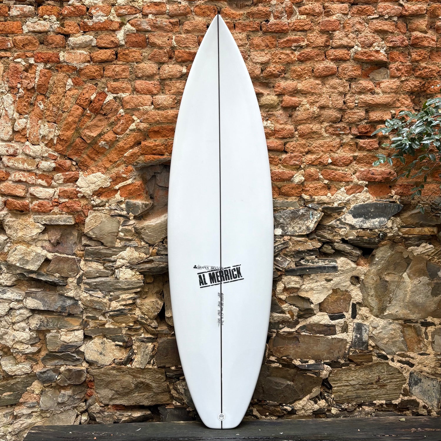 White CI 2.PRO  6’0	19 1/8	2 7/16	29.9L surfboard with 'Al Merrick' branding against a rustic stone wall.