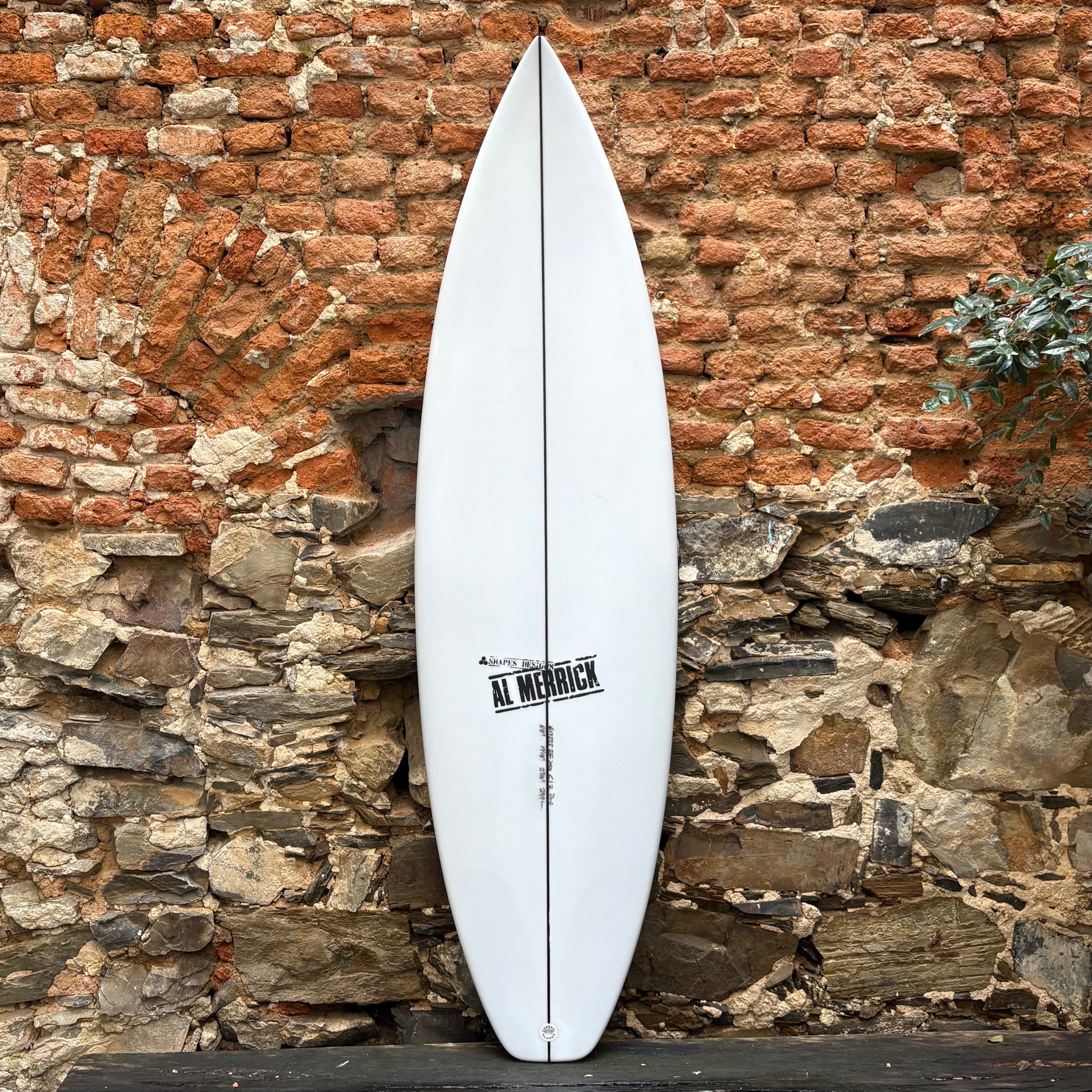White CI 2.PRO  6’0	19 1/8	2 7/16	29.9L surfboard with 'Al Merrick' branding against a rustic stone wall.