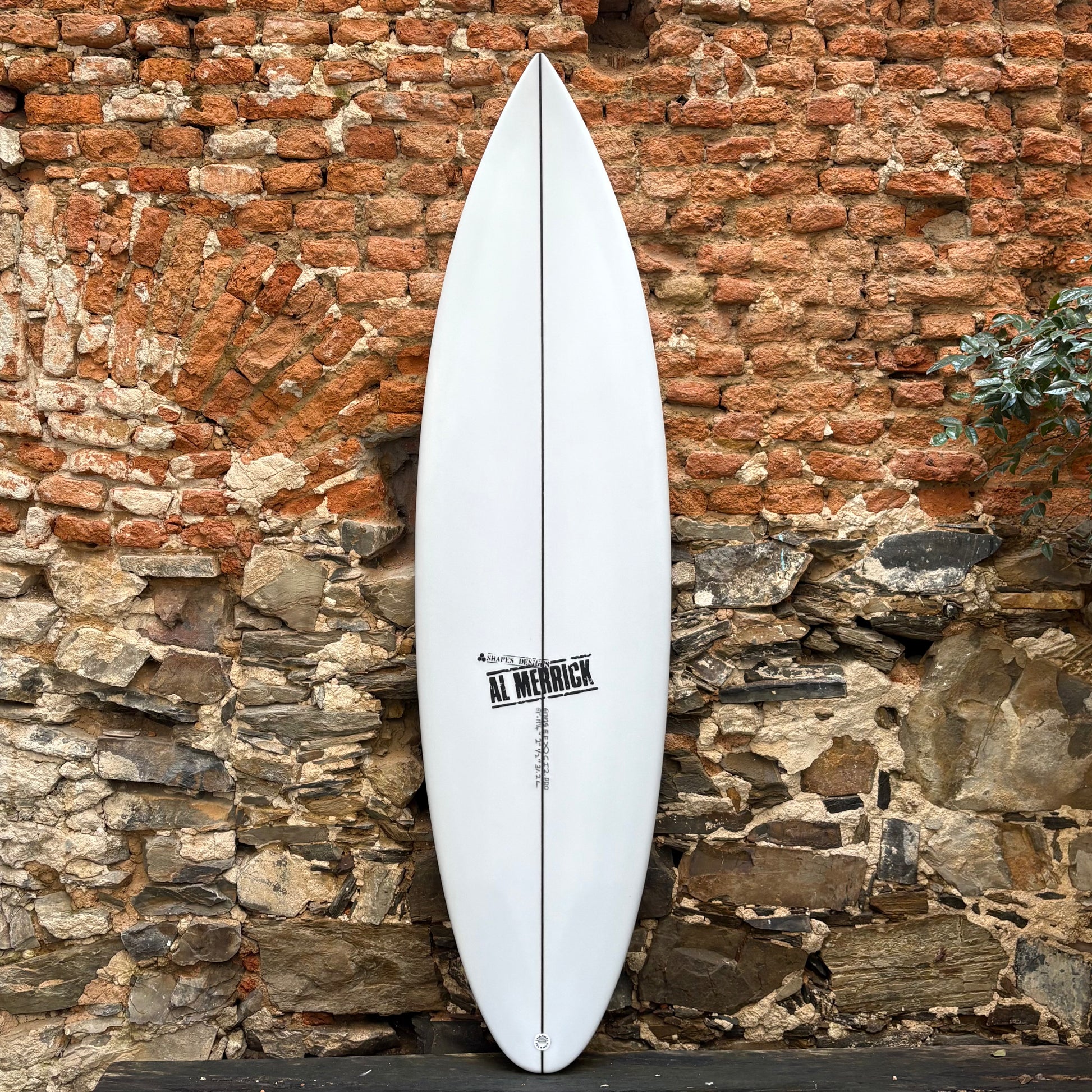 White CI 2.PRO  6’1	19 1/4	2 1/2	31.2L  surfboard with 'Al Merrick' branding against a rustic stone wall.