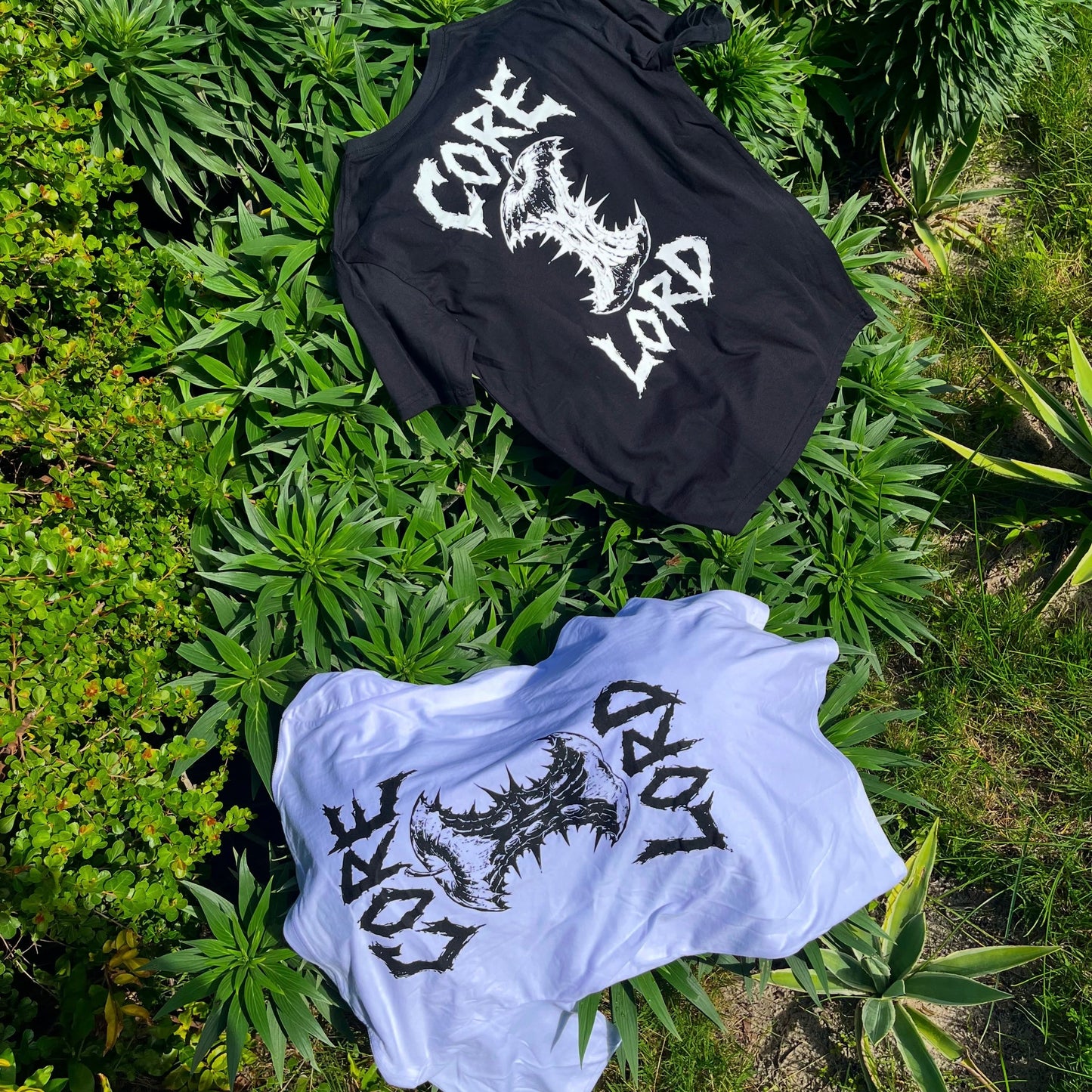 Two graphic t-shirts laid out on green foliage. The top black t-shirt features a white spiky logo reading 'CRAZE LORD', and the bottom light blue t-shirt features a black spiky logo reading 'CRAZE LORD' with a skull graphic.