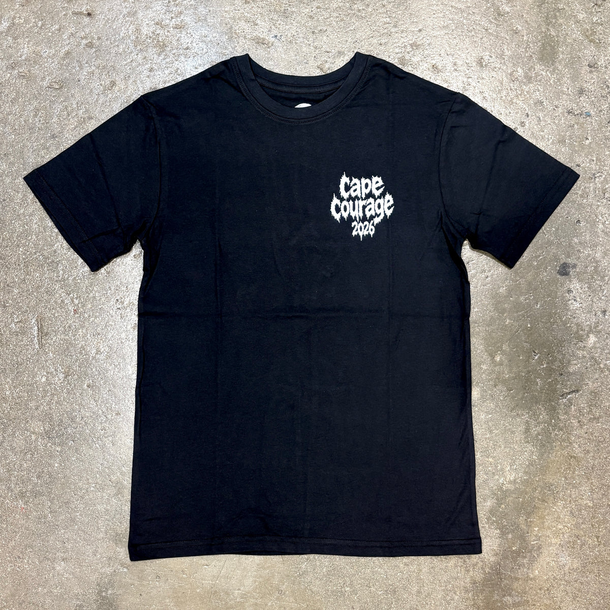 A black short-sleeve t-shirt with a white 'Cape Courage 2026' graphic printed on the left chest, laid flat on a textured concrete surface.