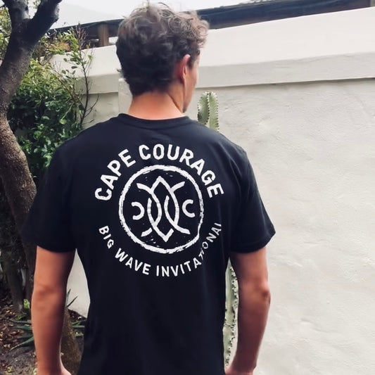 A man viewed from behind wearing a black t-shirt with a large white graphic on the back that reads 'CAPE COURAGE' and 'BIG WAVE INVITATIONAL'.