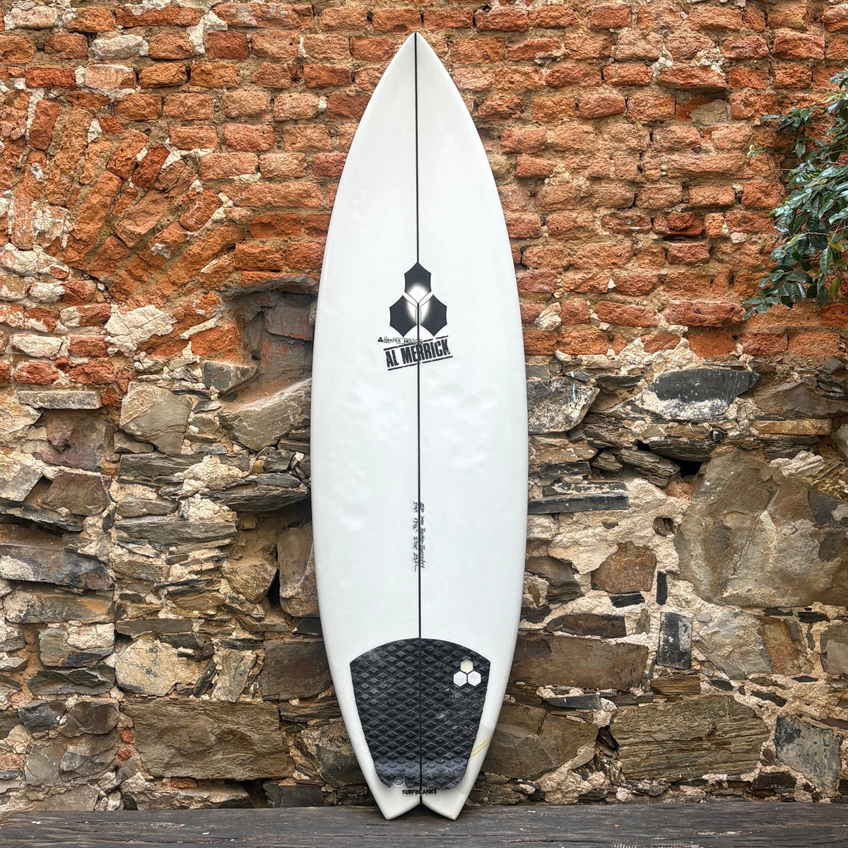 A white  surfboard with a black graphic logo and a black grip pad, leaning against a rustic brick and stone wall.