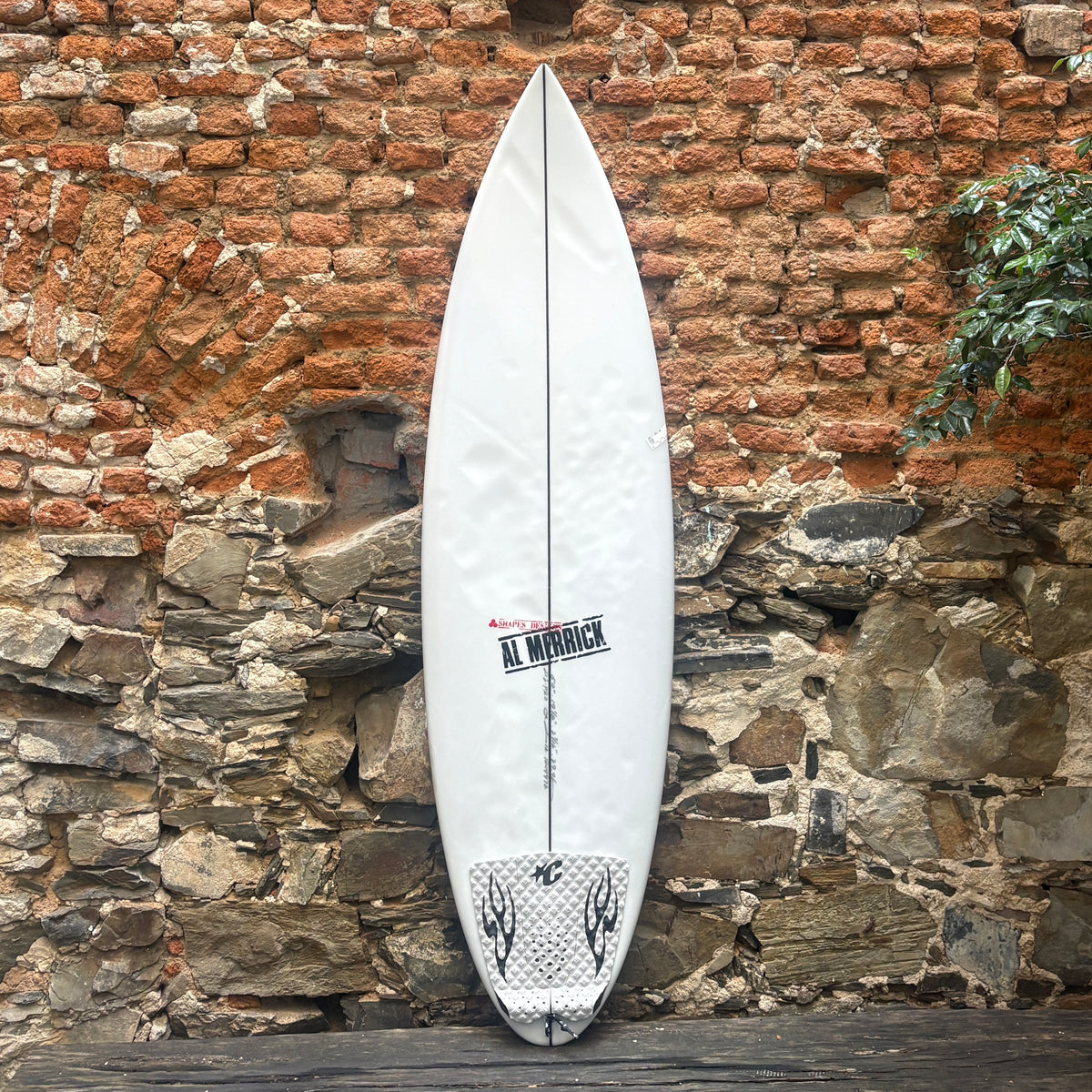 A white surfboard with black graphics and the Al Minnick brand logo, leaning against a rustic stone and brick wall.